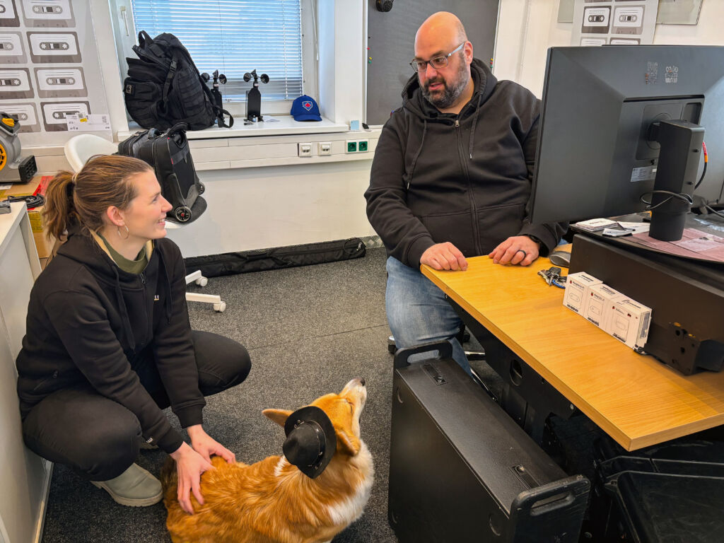 Zwei Kollegen, die sich an einem Schreibtisch unterhalten; ein Hund sitzt zwischen ihnen.