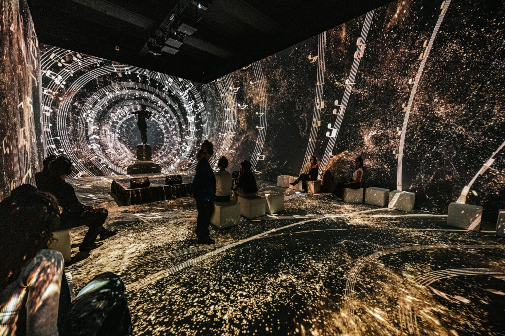 Projektionen aus goldenen Noten und Funken füllen einen 360-Grad-Erlebnisraum; Personen beobachten das Schauspiel.