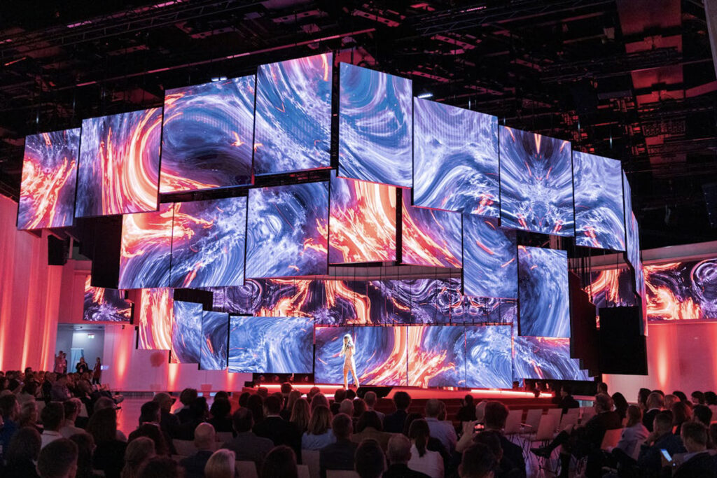 Eine Bühne, über der zahlreiche quadratische LED-Displays in unterschiedlicher Anordnung schweben. Darauf sind ineinander fließende Inhalte aus blauen und orangen Wellen und Kreisen zu sehen. Davor ein Publikum, das die Geigerin untern den LED-Displays auf der Bühne bestaunt.